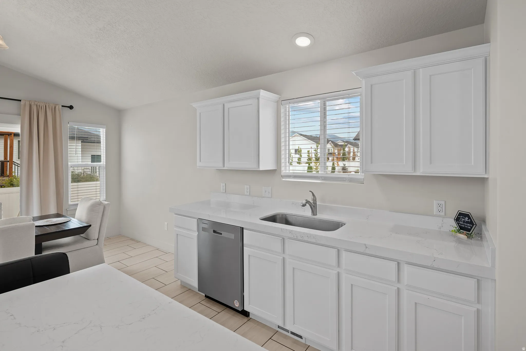White shaker cabinetry, light-colored countertops, stainless steel dishwasher, undermount sink with a chrome faucet, and tiled flooring