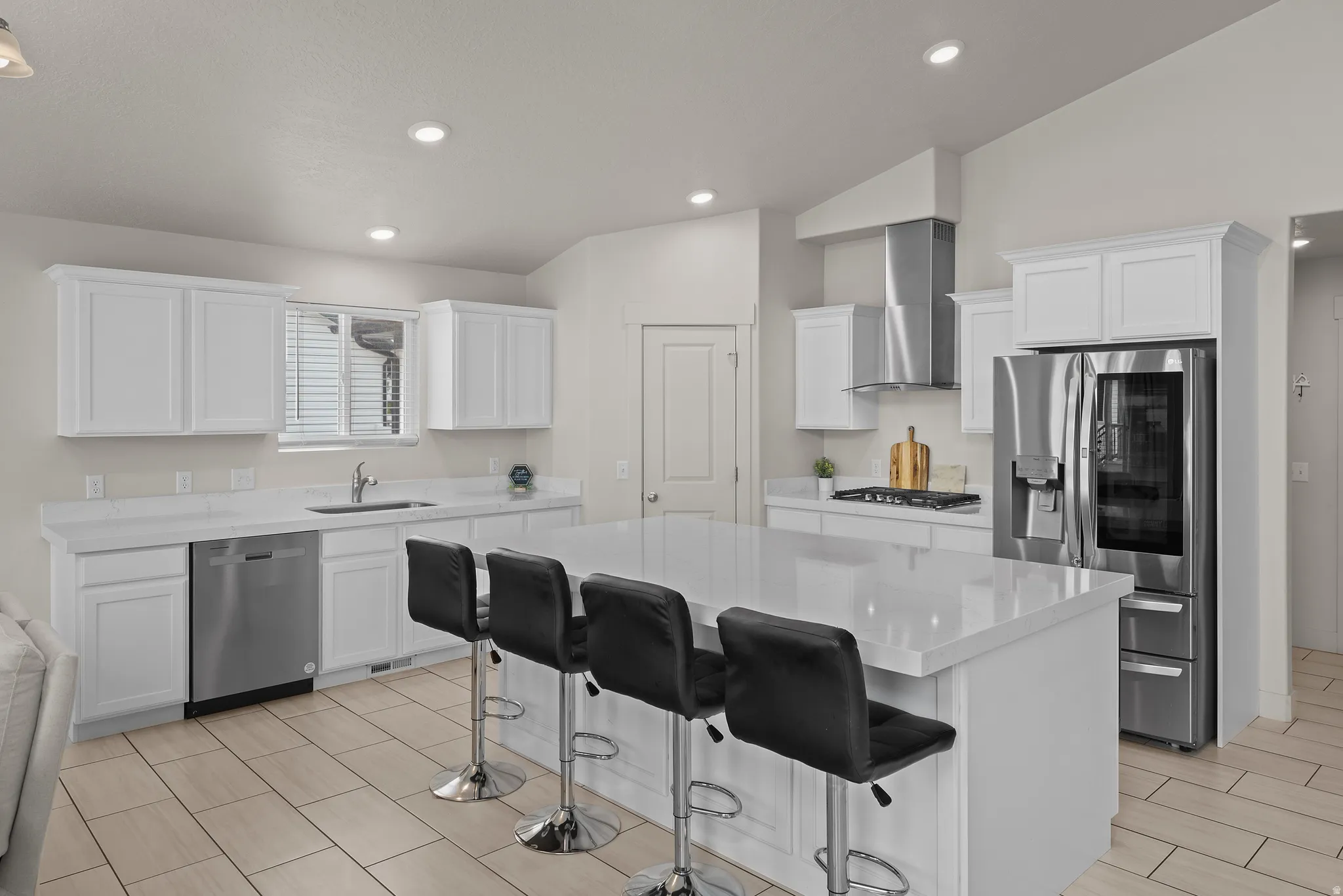 Modern kitchen featuring white shaker-style cabinetry, stainless steel appliances including a built-in refrigerator and range hood, quartz countertops, a large island with seating for four, tile flooring, and recessed ceiling lighting throughout