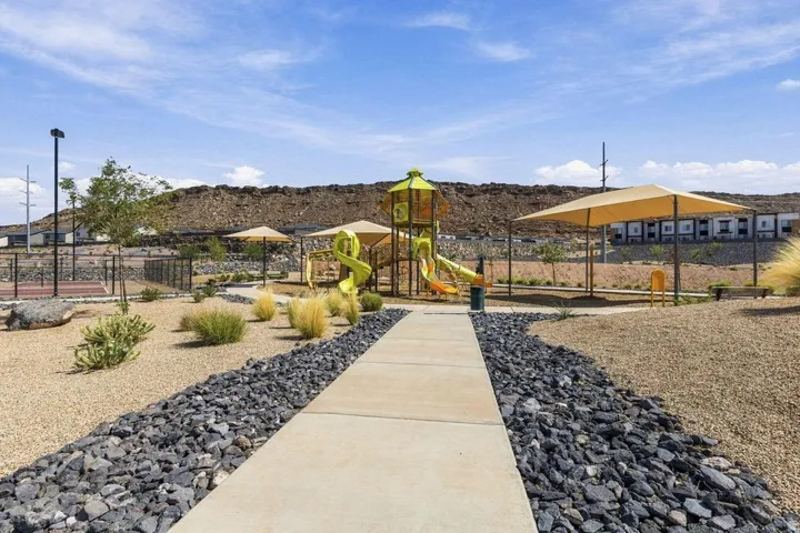 Playground structure with slides and climbing features, shaded seating areas, concrete pathways, decorative rock landscaping, and a fenced court