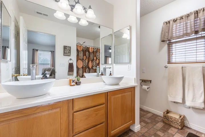 Dual vessel sinks, light wood-finish vanity with storage, expansive wall mirror, overhead vanity lighting, and a window with horizontal blinds