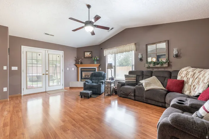 Vaulted ceiling with a ceiling fan, wood-finish flooring, white French doors, corner fireplace with a wood mantle, and a large window
