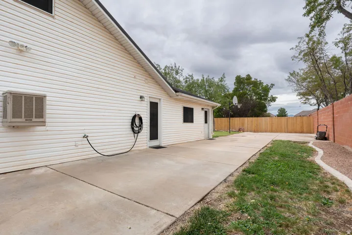 Paved concrete patio area extending along the exterior, white vinyl siding, a rear entry door, and perimeter fencing