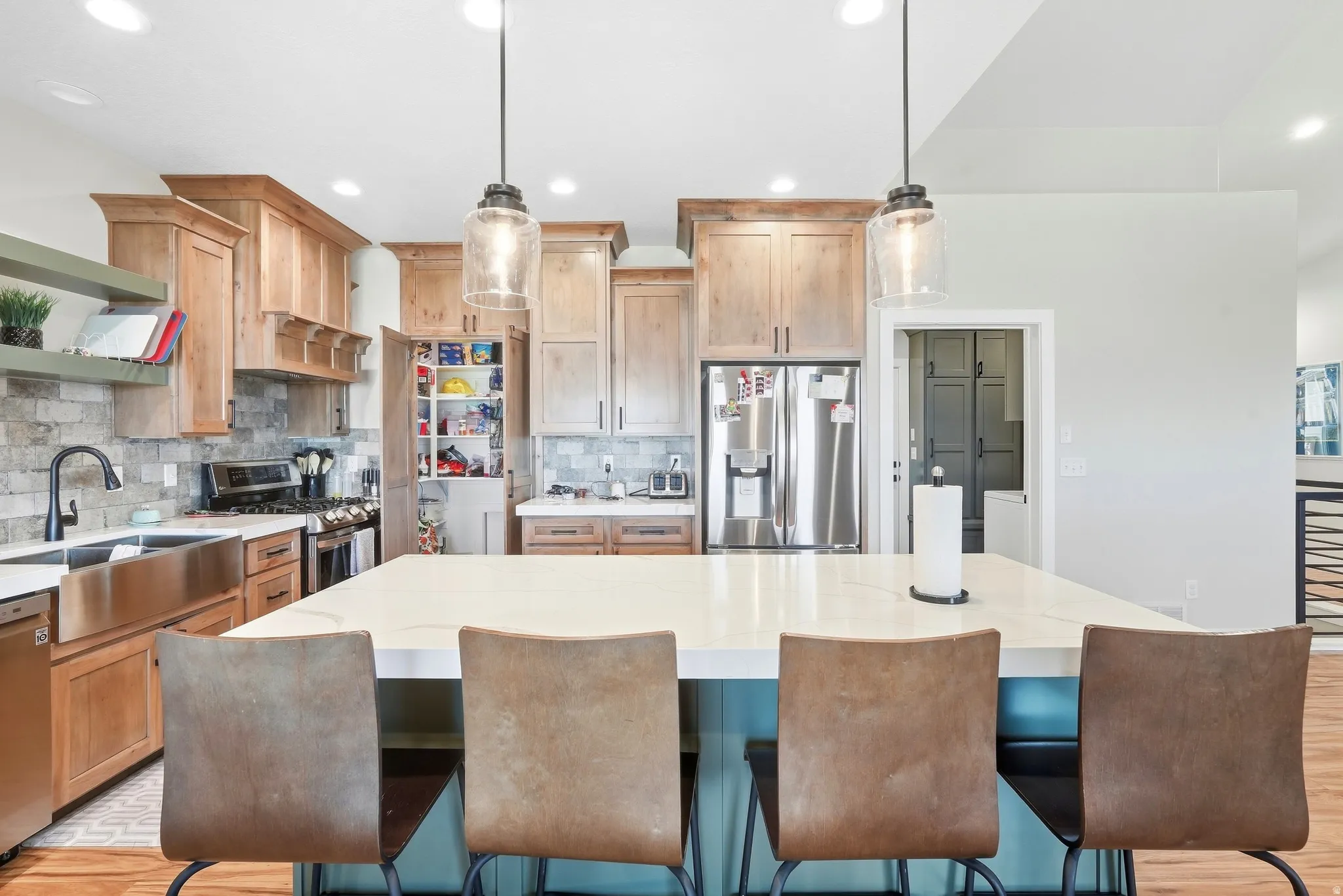Kitchen featuring a large center island with a light-toned countertop, stainless steel appliances, stone-finish backsplash, wood cabinetry, and a stainless steel farmhouse sink