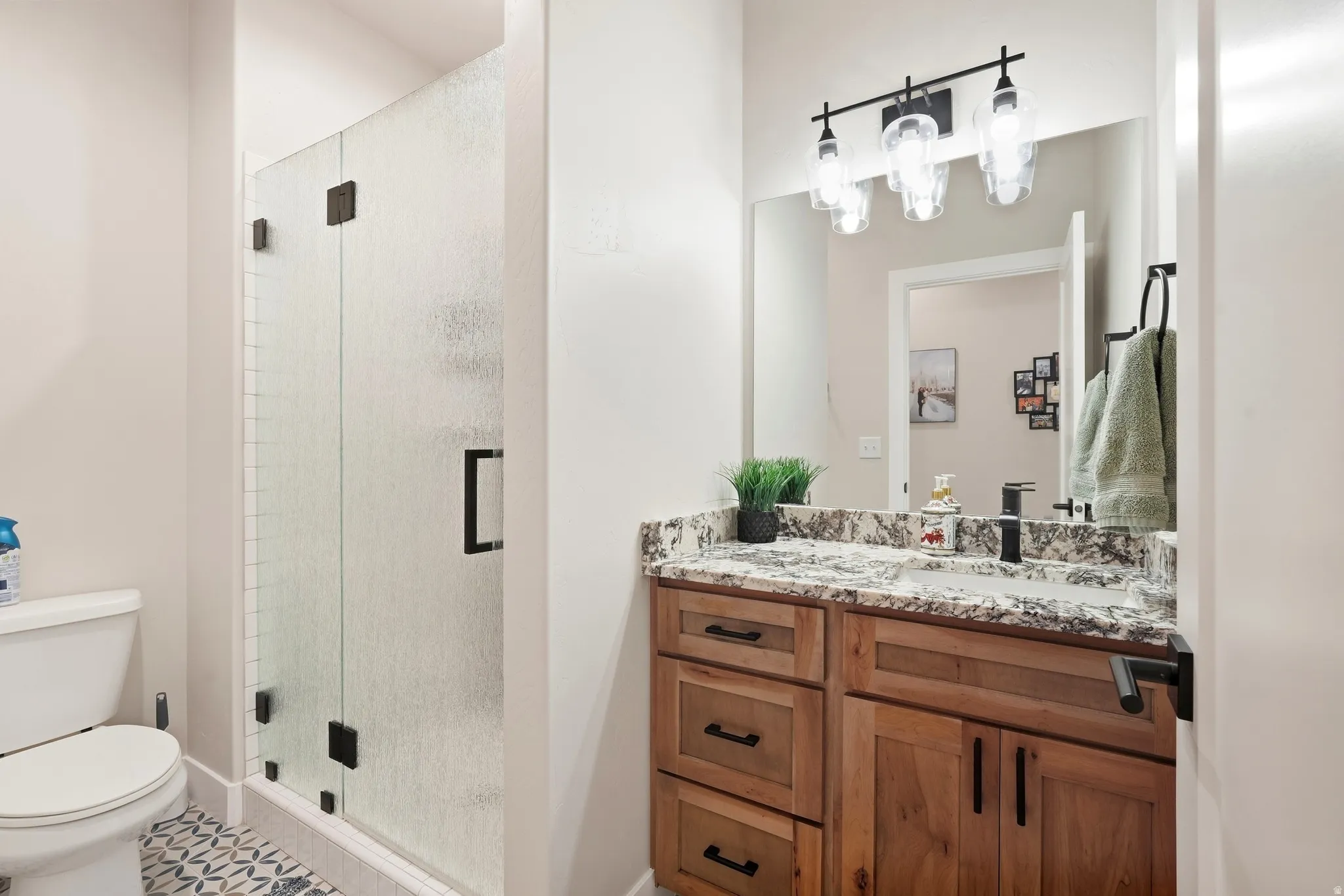 Walk-in shower with frosted glass enclosure, dual vanity with stone countertop, wood-finish cabinetry, and patterned floor tiling