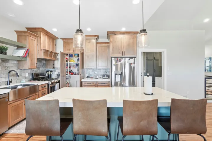 Kitchen featuring a large center island with a light-toned countertop, stainless steel appliances, stone-finish backsplash, wood cabinetry, and a stainless steel farmhouse sink