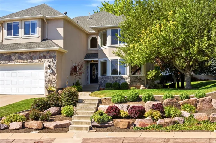 Stucco exterior with stone accents, attached garage, multi-level landscaping with mature trees, and a concrete walkway