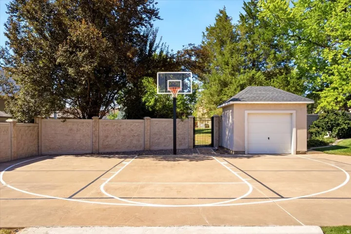 Outdoor sports court with painted lines, a basketball hoop, stucco perimeter wall, and a detached single-car garage with an asphalt shingle roof