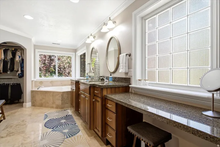 Dual vanity with wood cabinetry and stone countertops, a soaking tub with tile surround, a built-in dressing table, and a glass block window