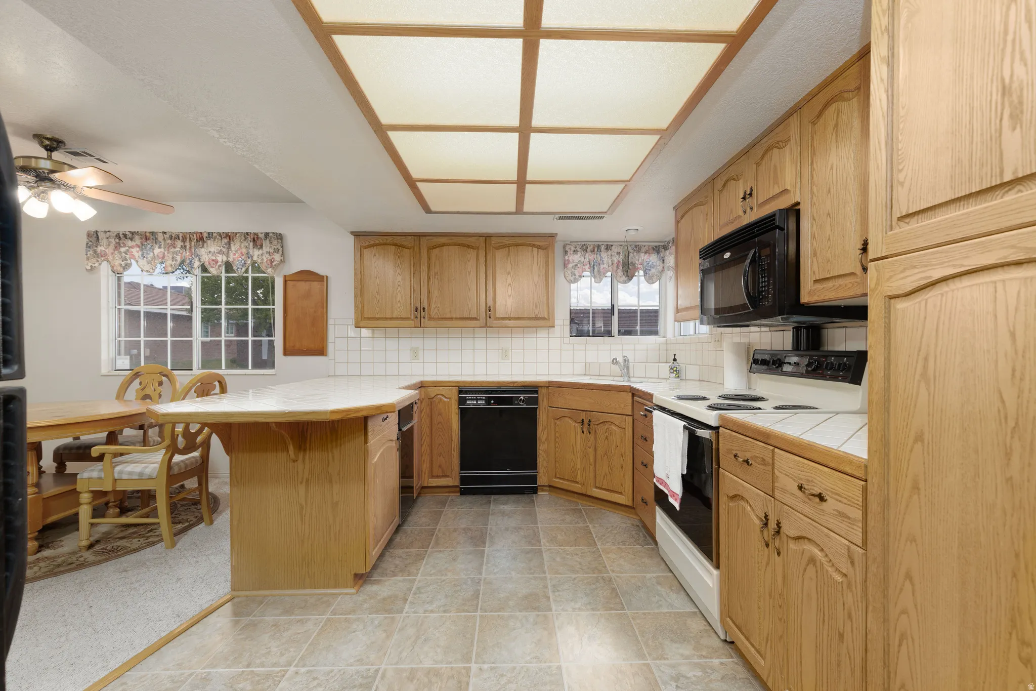 Kitchen featuring wood cabinetry, white tile countertops, black appliances, a tile backsplash, and a breakfast bar