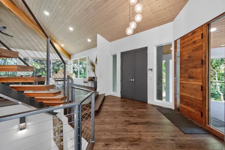 Hardwood flooring throughout the entry, exposed wood ceiling, modern floating staircase with cable railing, large windows, and a solid wood entry door