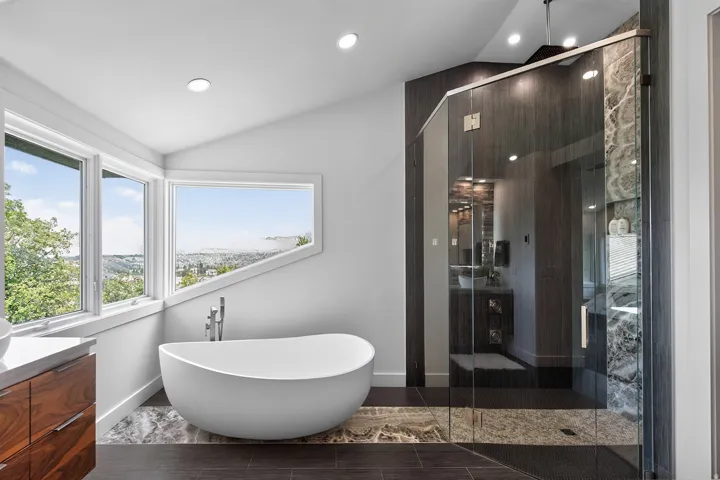 Freestanding soaking tub, multiple windows, glass-enclosed shower with tiled accents, wood-finish vanity with countertop, and recessed lighting