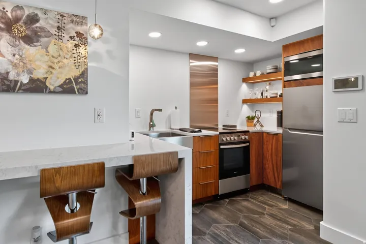 Kitchen featuring a breakfast bar with stone countertop, stainless steel appliances, wood-finish cabinetry, open shelving with integrated lighting, and hexagonal tile flooring