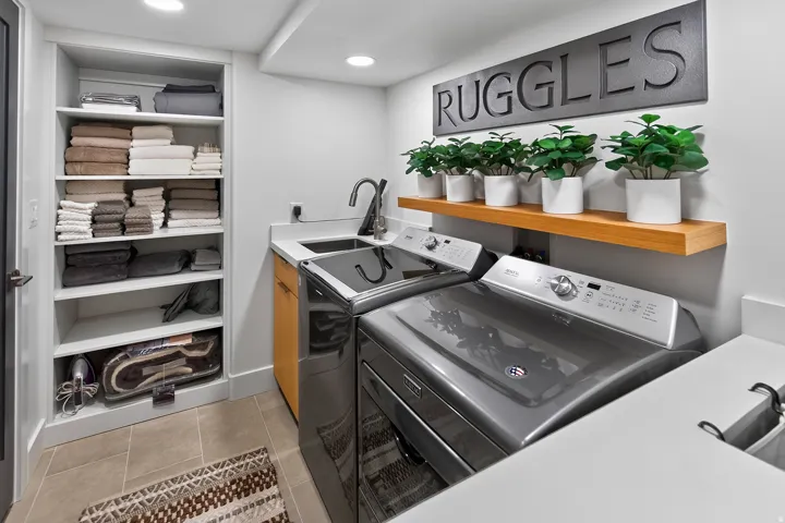 Integrated laundry area featuring a built-in shelving unit, stone countertop, undermount sink with a gooseneck faucet, and a wood-finish floating shelf