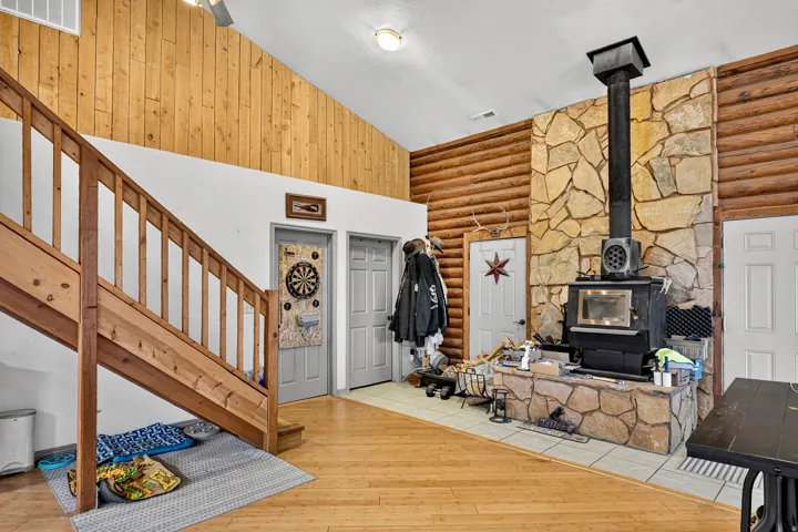 Natural wood flooring, stone-clad fireplace with wood-burning stove, exposed wood beam ceiling, natural wood paneling, and a natural wood staircase