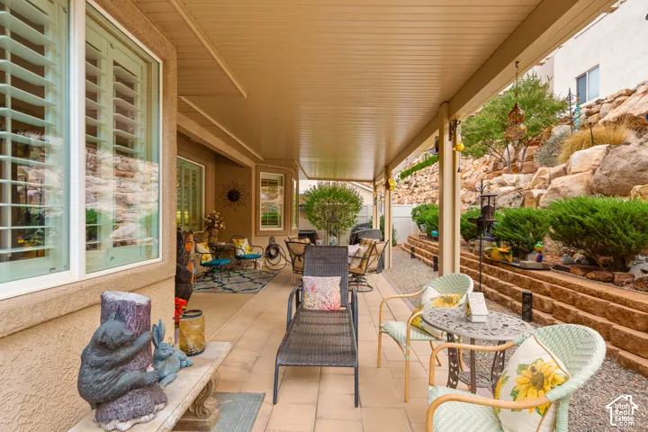 Covered patio with a paver surface, white framed windows, and rock landscaping with retaining walls