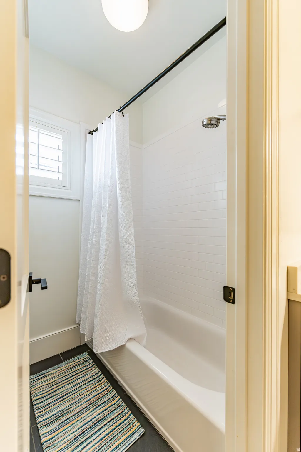 Tub and shower combination with white subway tile surround, matte black shower rod, and a window with white louvered shutters