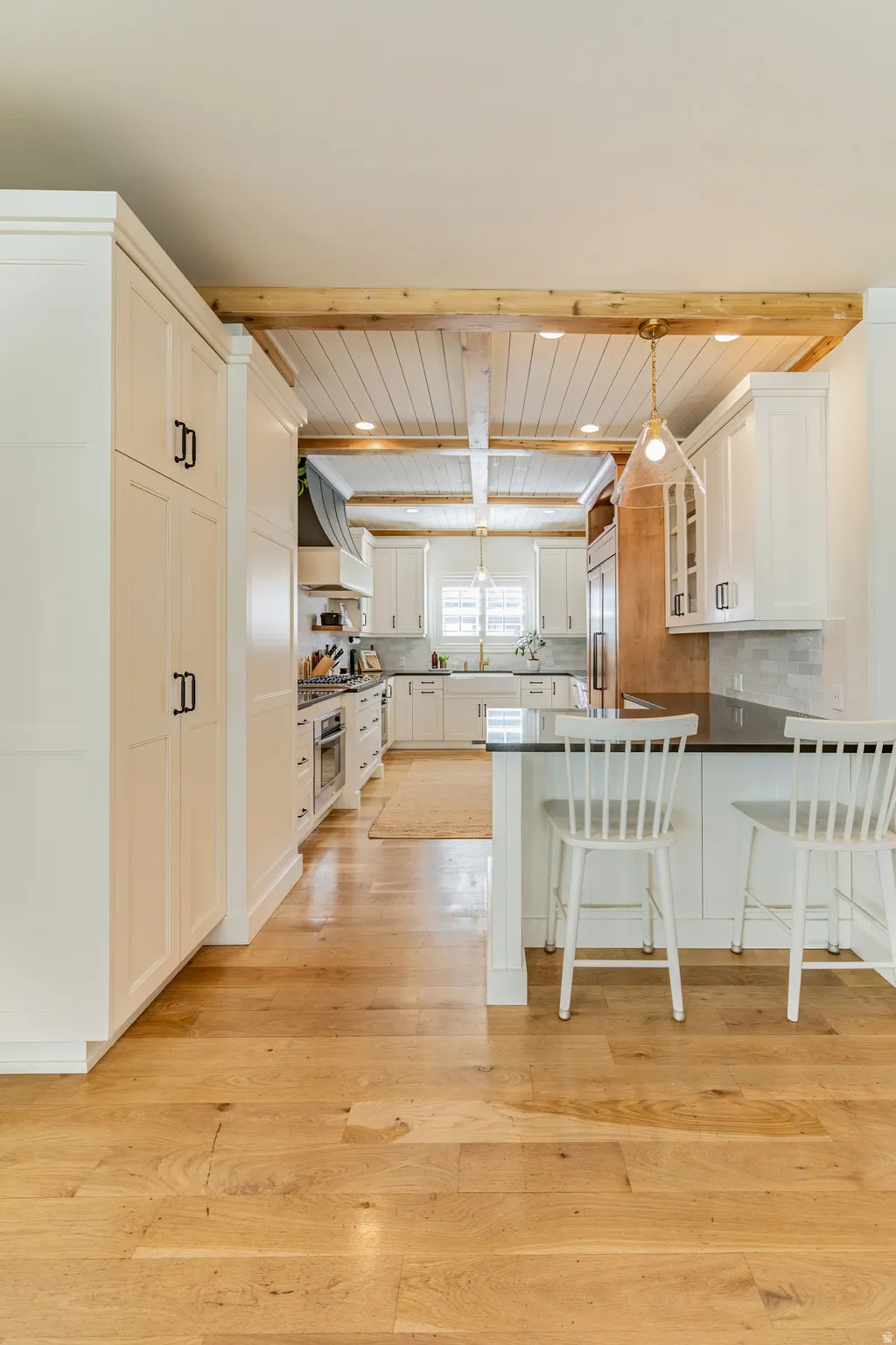 White shaker cabinetry, wood-finish flooring, exposed wood beams, black countertops, and a white subway tile backsplash
