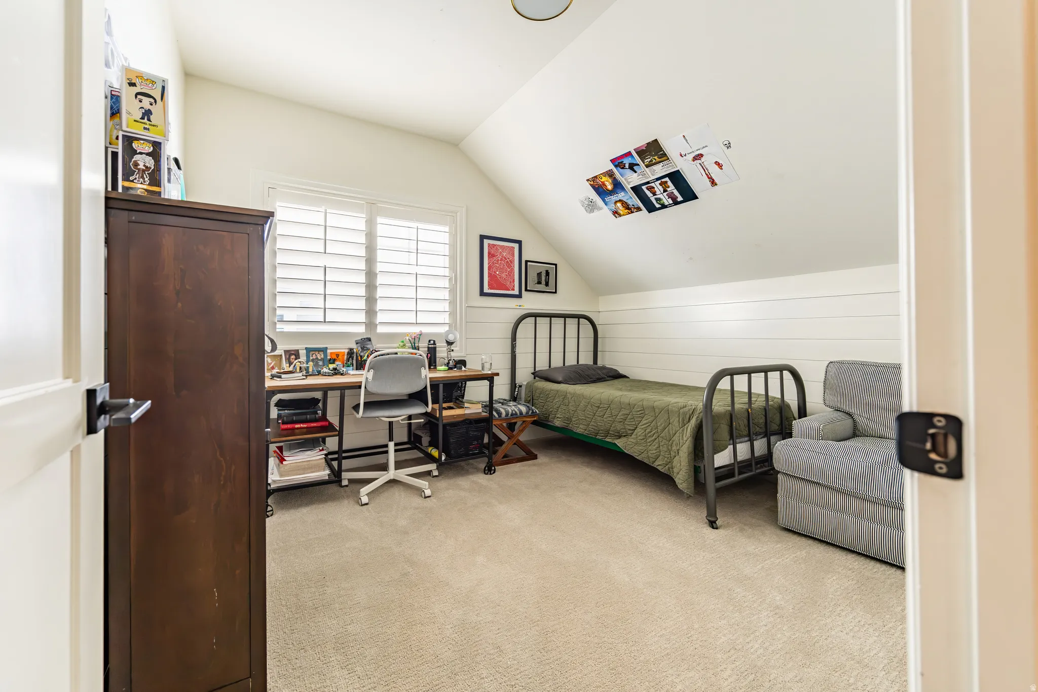 Sloped ceiling, neutral-toned carpeting, window with plantation shutters, white paneled wall accent, and a black door handle