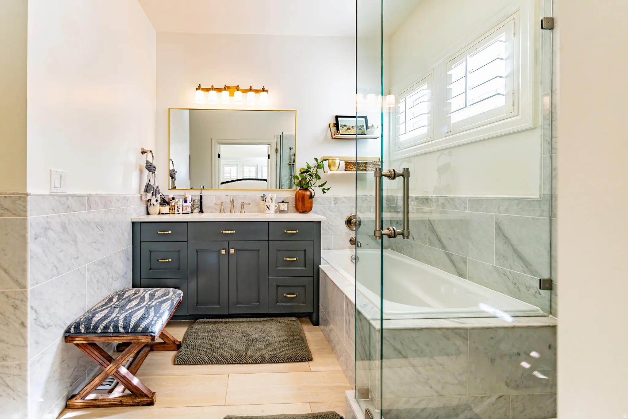 Dual sink vanity with gold-tone hardware, full-height marble tile surround, built-in bathtub, and glass-enclosed shower