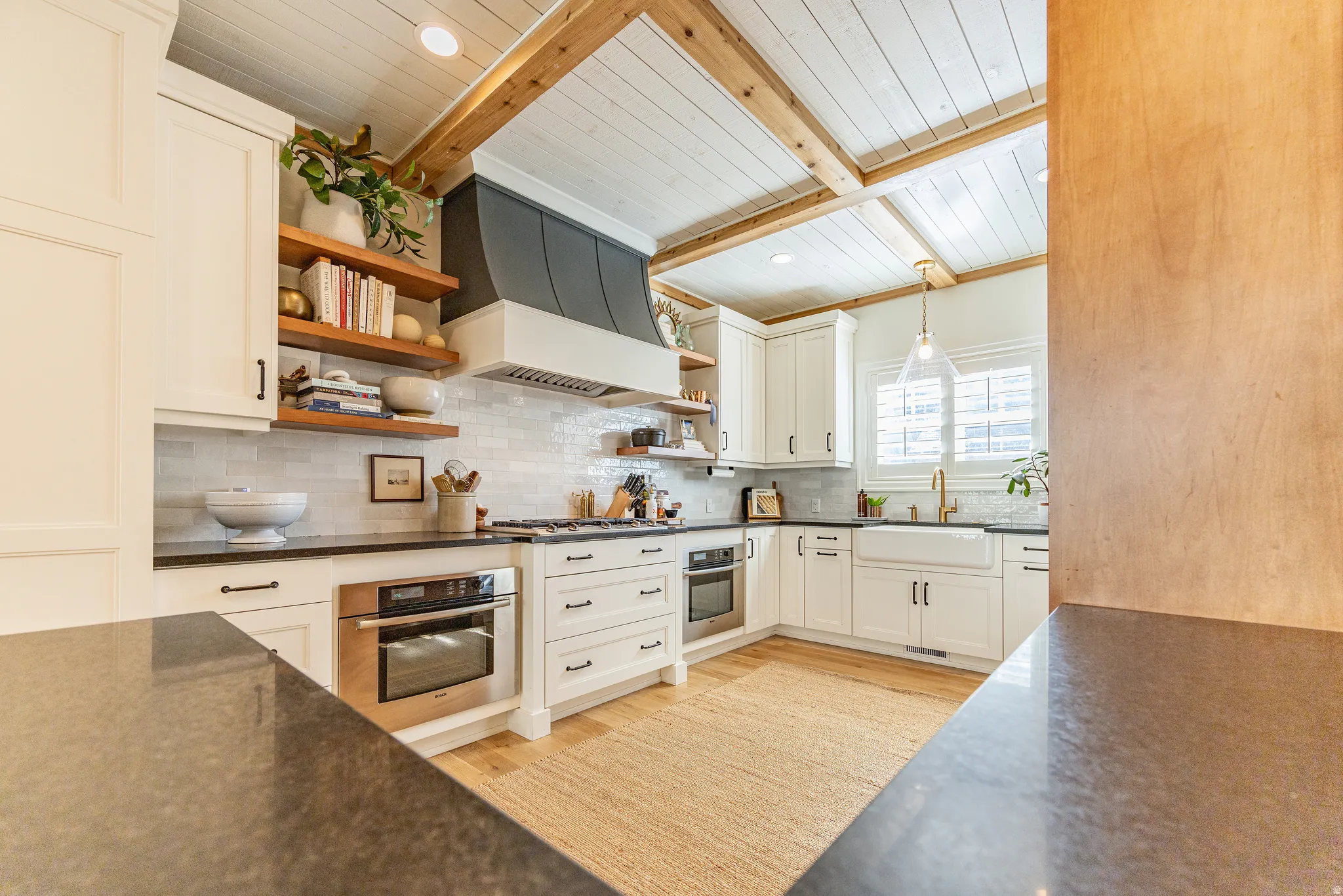 White shaker cabinetry, black countertops, stainless steel appliances, exposed wood beam ceiling, and a white farmhouse sink
