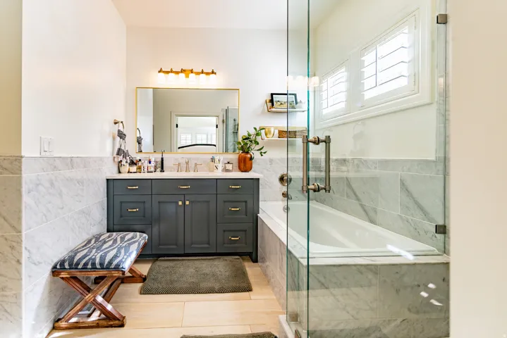 Dual sink vanity with gold-tone hardware, full-height marble tile surround, built-in bathtub, and glass-enclosed shower