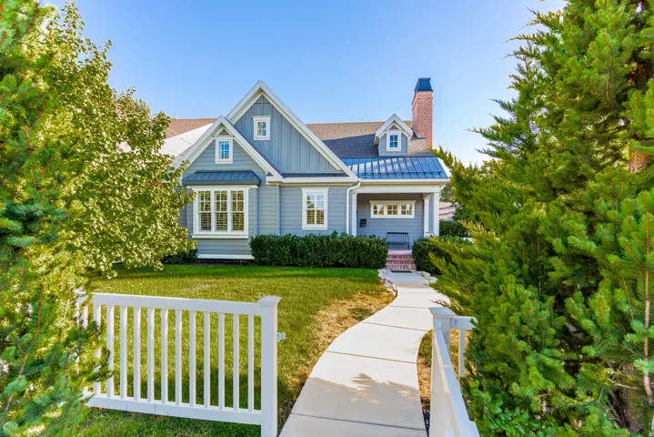 Light blue siding with white trim, bay window, front porch with columns, brick chimney, and a white picket fence