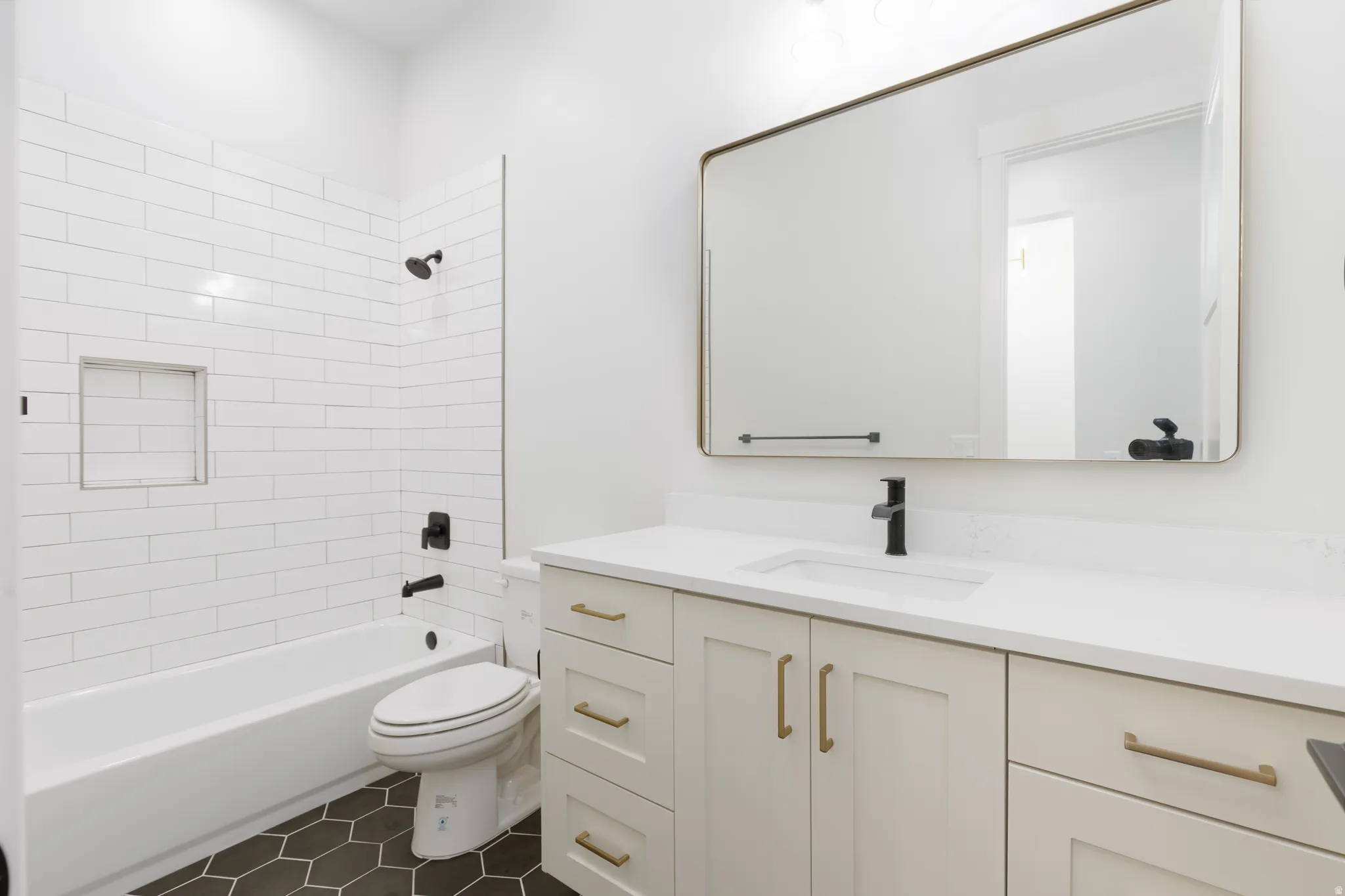 White subway tile shower surround with integrated niche, dark hexagon tile flooring, light-toned vanity with gold-finish hardware, white countertop with integrated sink, and a rectangular mirror with gold-finish frame