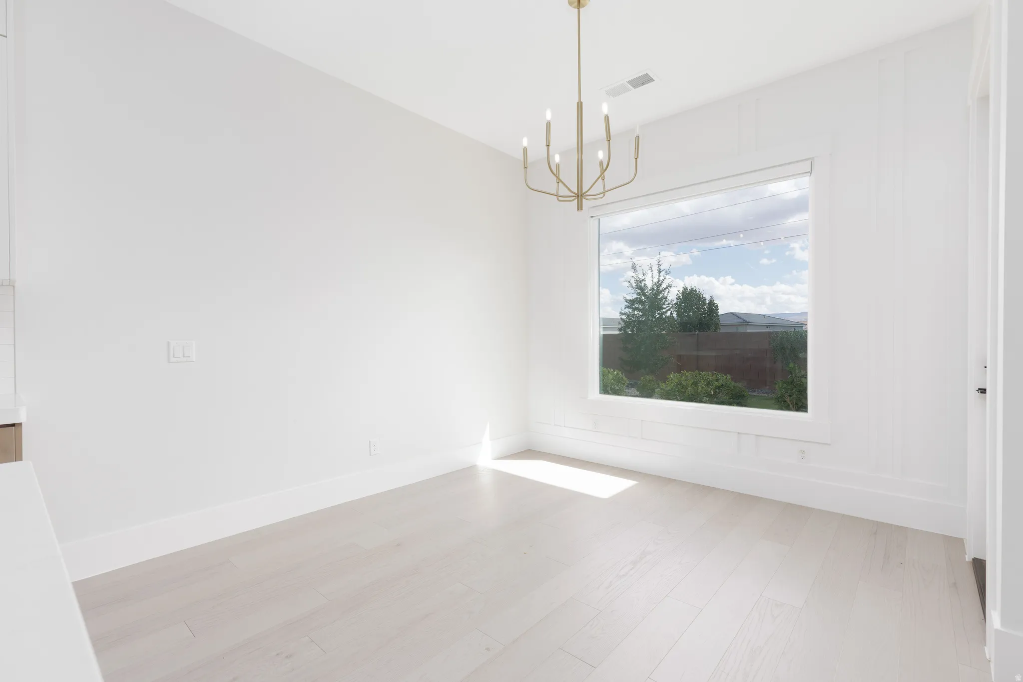 Wide plank wood-finish flooring, large picture window, board and batten wall accent, contemporary chandelier, and bright white walls