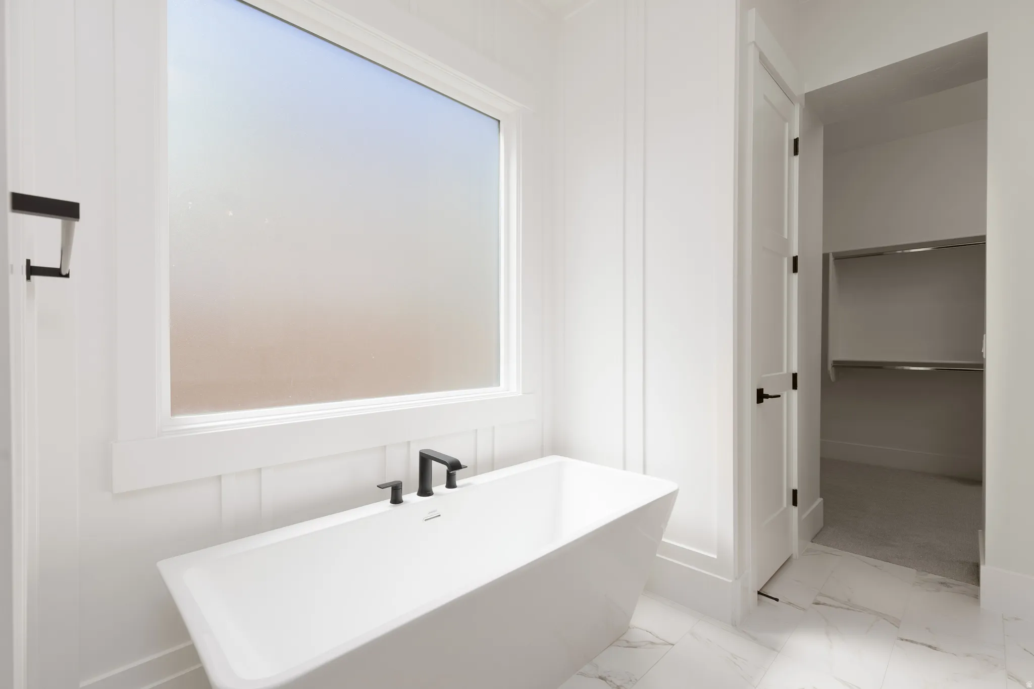 Freestanding soaking tub with matte black fixtures, frosted window, white paneling, and marble-look floor tiling