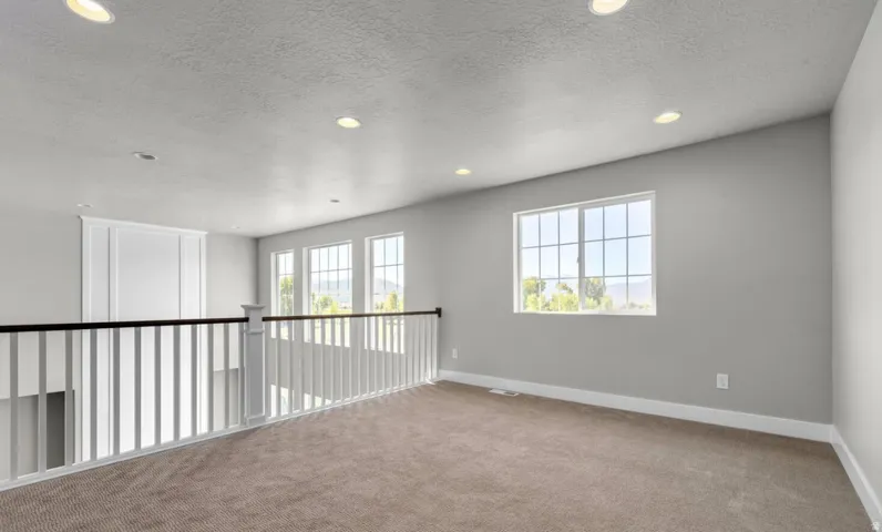 Recessed lighting, light gray wall finishes, light brown carpeting, multiple windows, and a white and black spindle railing