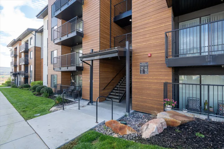 Multi-story building featuring wood-finish siding, black metal railings, concrete walkways, and landscaped grounds with natural rock accents