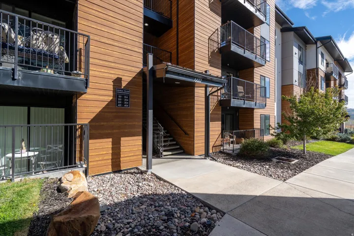 Exterior featuring wood-look siding, black metal railings, concrete walkways, a landscaped planting bed with small trees, and a ground-level patio