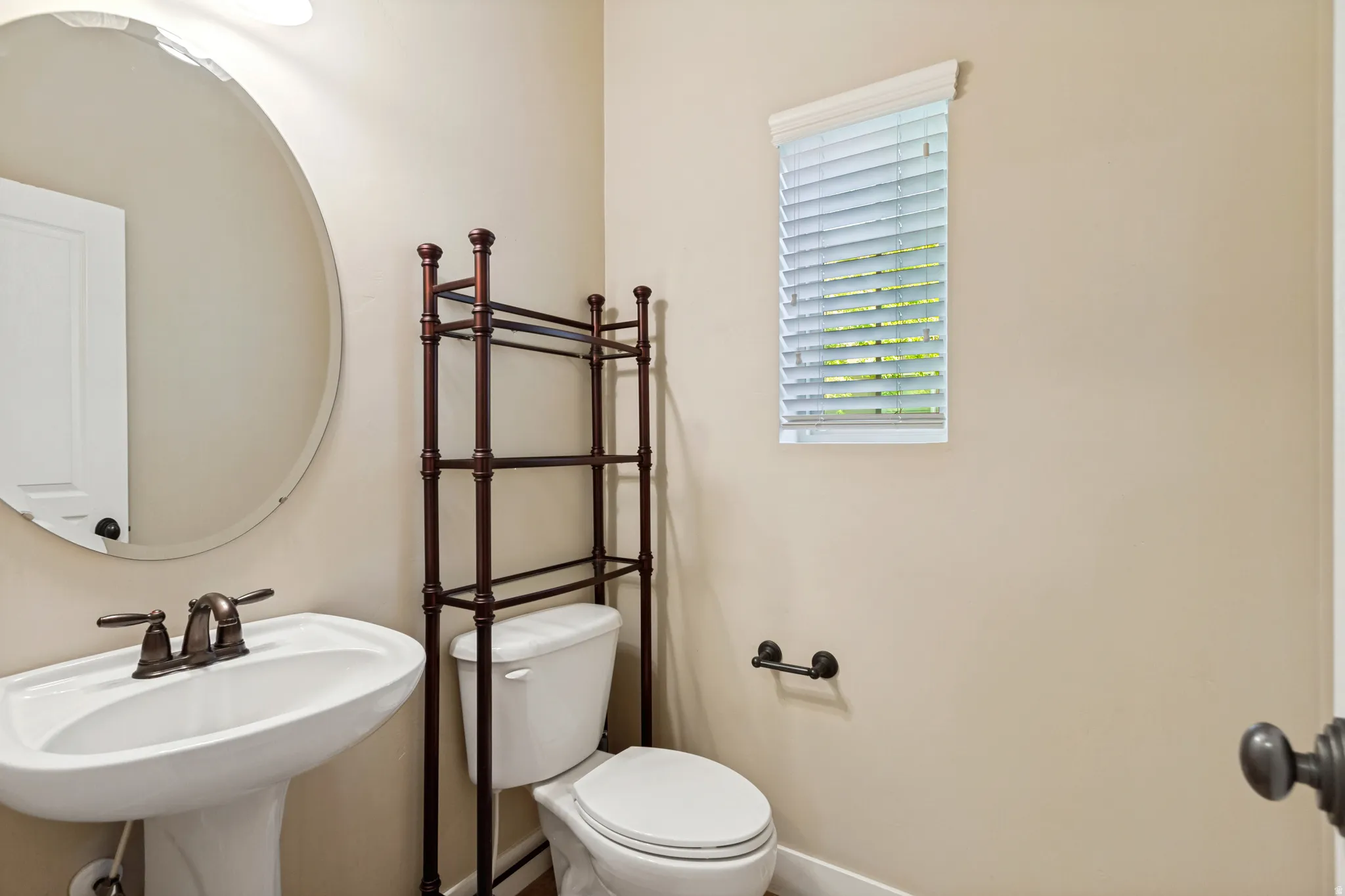 Pedestal sink with bronze-finish faucet, oval mirror, toilet, and window with blinds