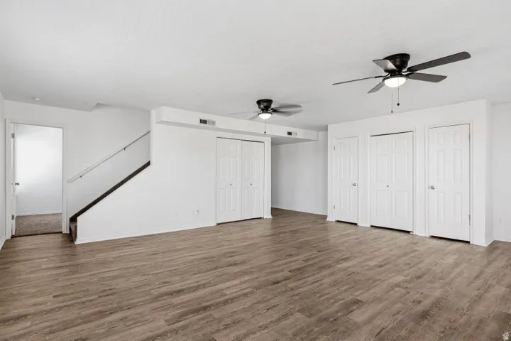 Wood-finish flooring, white walls, multiple ceiling fans, staircase with dark handrail, and several closed storage doors