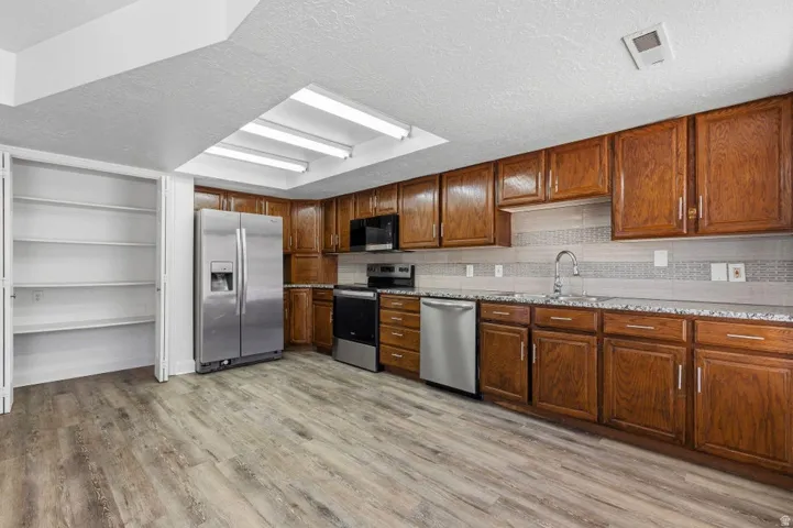 Spacious kitchen featuring wood-finish flooring, wood cabinetry, stone-finish countertops, stainless steel appliances, and a tiled backsplash