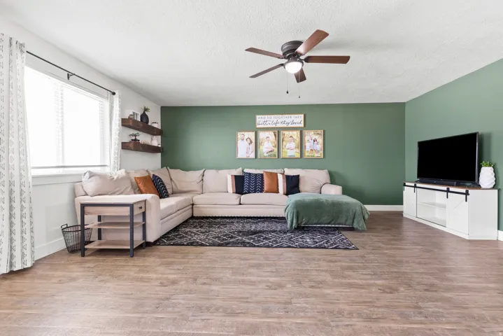 Wood-finish flooring, accent wall, ceiling fan with integrated lighting, and a window with blinds
