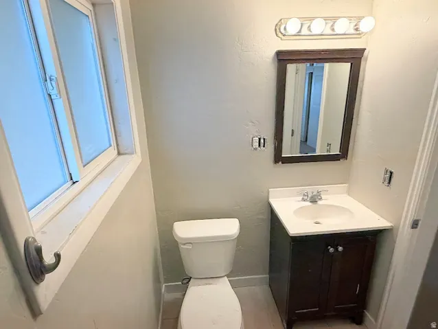 Compact bathroom featuring a window, toilet, dark wood-finish vanity with a white countertop and integrated sink, and a framed mirror with overhead lighting