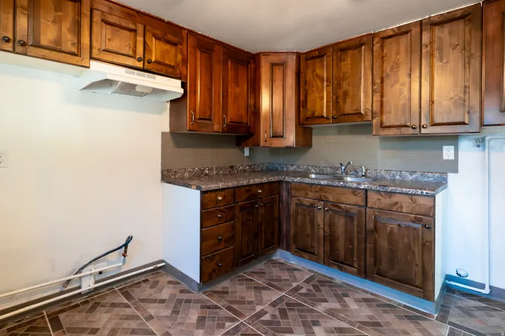 Wood cabinetry with dark hardware, stone-look countertops, stainless steel sink, subway tile backsplash, and patterned tile flooring