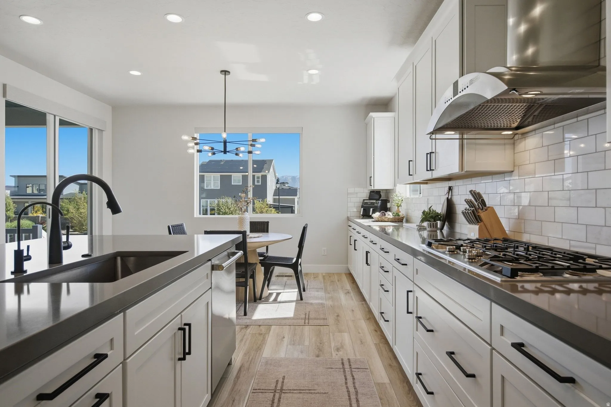 Modern kitchen featuring light cabinetry, dark countertops, a stainless steel range hood, a tile backsplash, gas stove top and LVP wood-finish flooring.