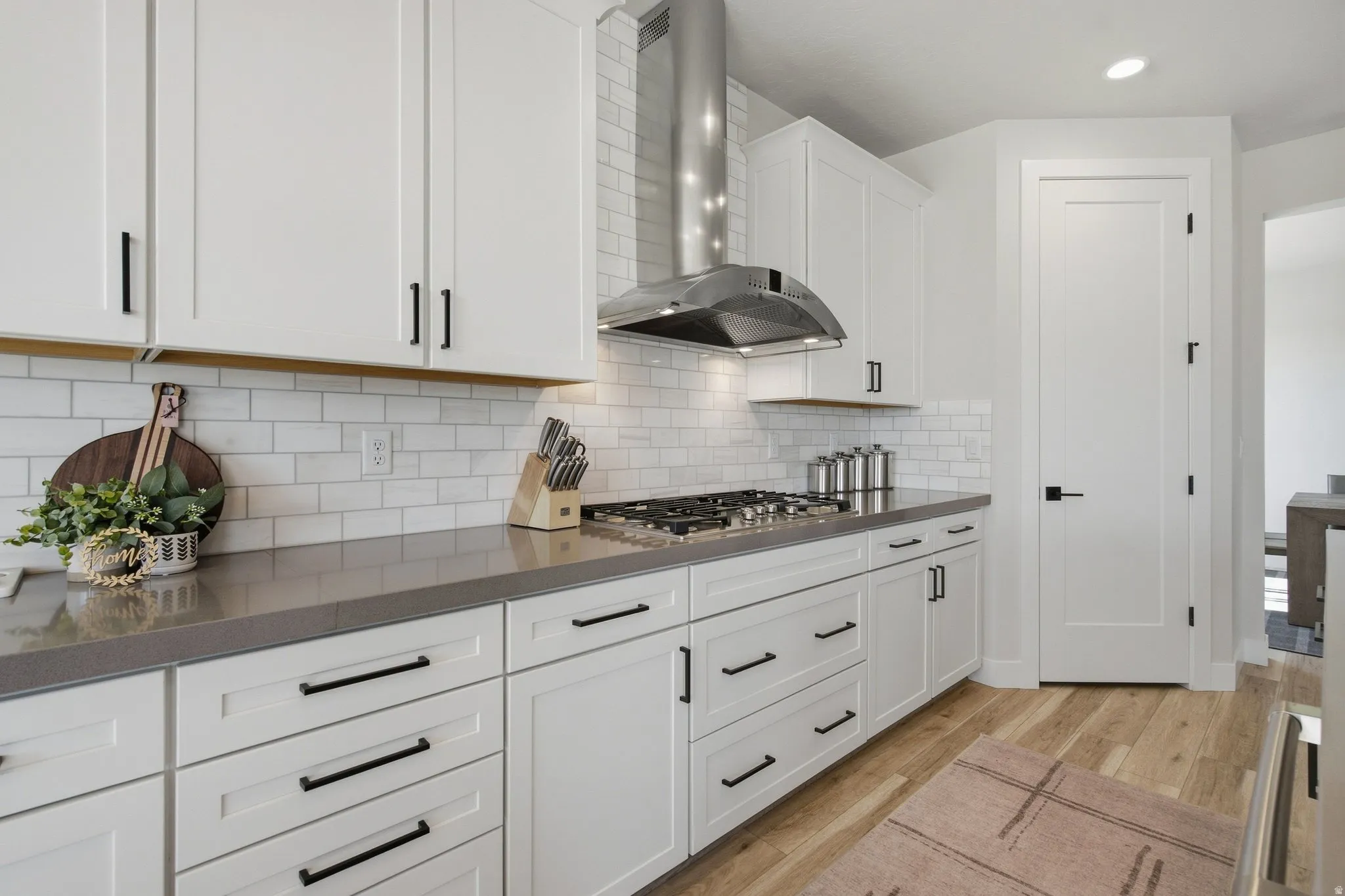 White cabinetry with matte black hardware, subway tile backsplash, dark-toned countertops, stainless steel range hood, and LVP wood-finish flooring.
