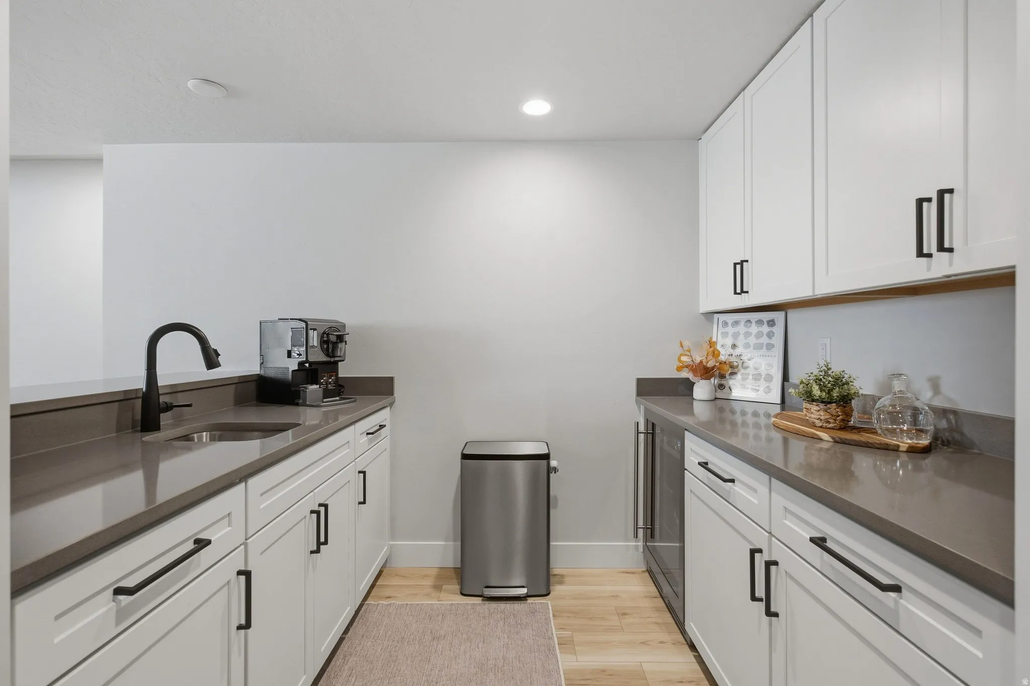 Wet bar with white cabinetry, dark countertops, stainless steel sink with a matte black faucet, and light LVP wood-finish flooring.