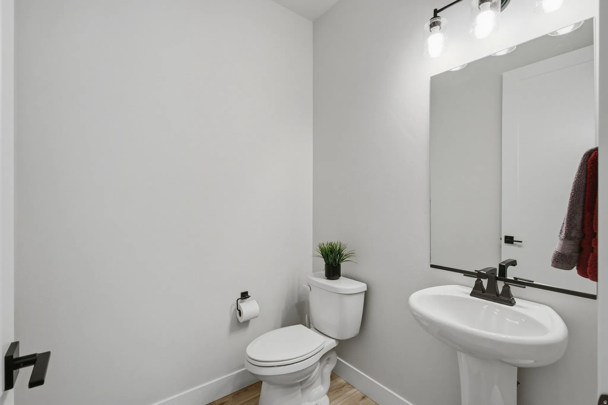 Half bath with pedestal sink with a dark faucet, wall-mounted mirror, and overhead lighting.