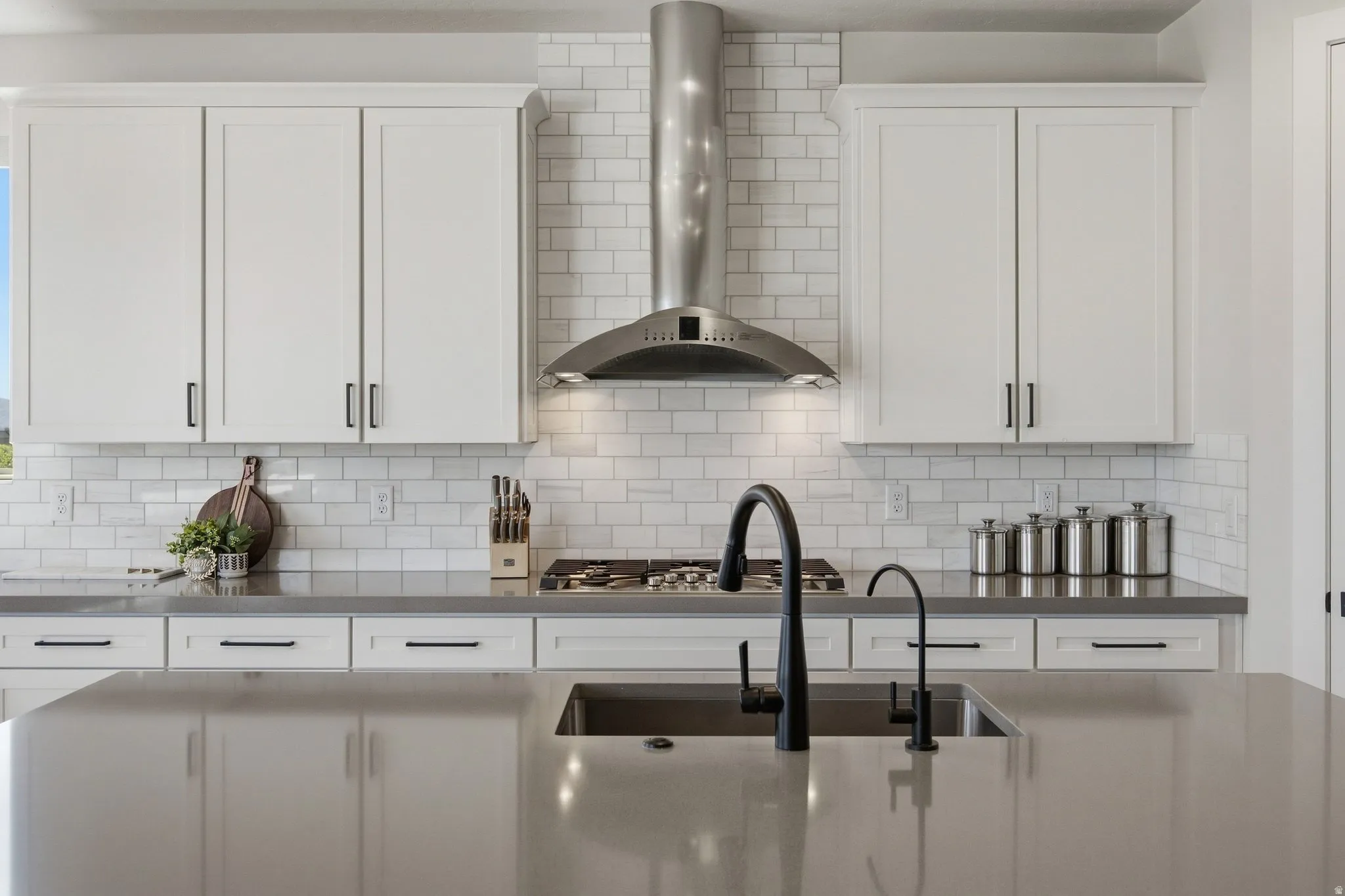 White cabinetry with matte black hardware, subway tile backsplash, stainless steel range hood, built-in gas range, and a large kitchen island with an undermount sink and matte black faucet.