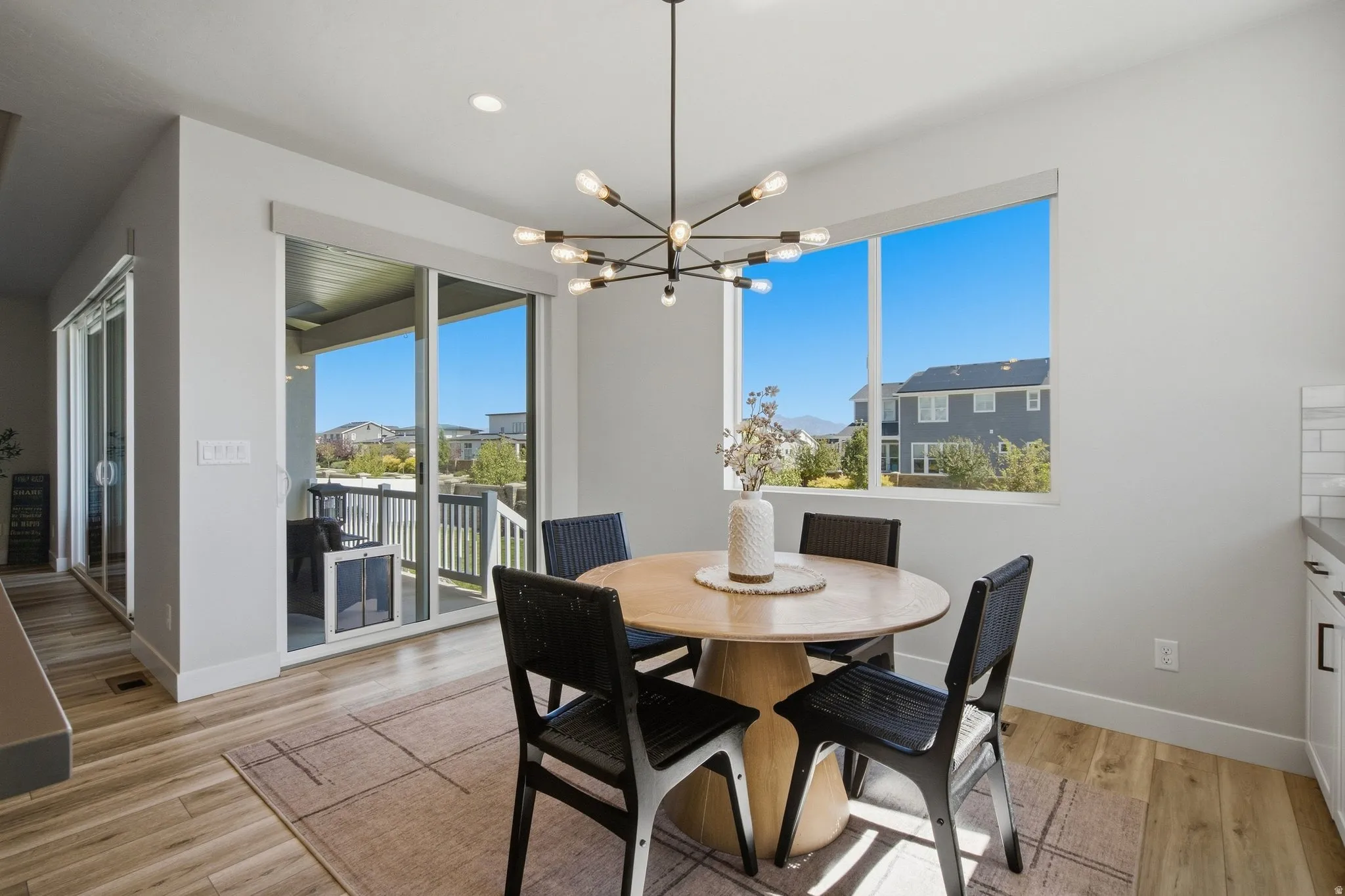 Spacious eating area featuring LVP wood-finish flooring, a contemporary chandelier, a large picture window, and sliding glass doors opening to an exterior covered patio with railings.