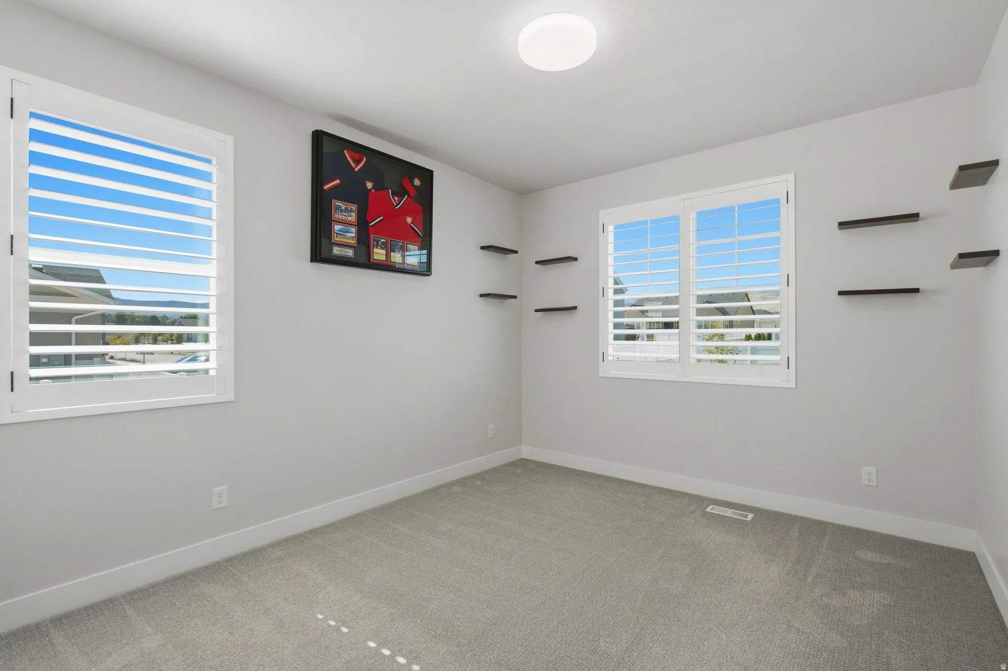 Light walls with white trim, two windows with white shutters, neutral carpet flooring, and dark floating shelves.