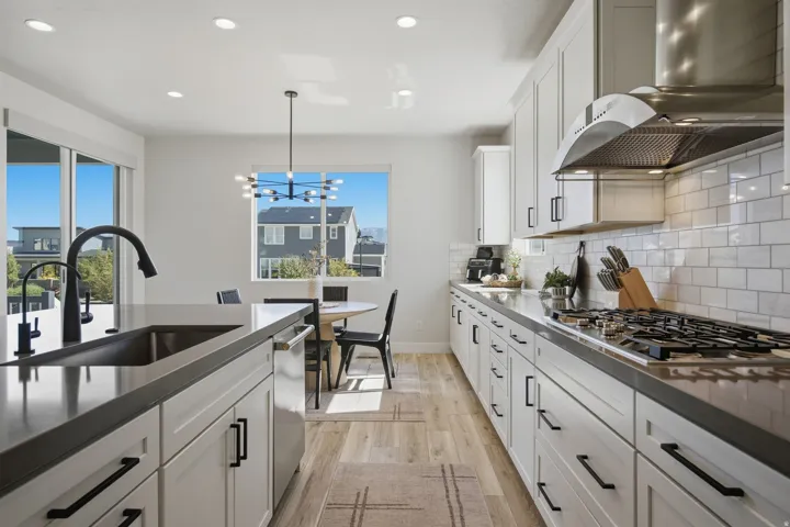 Modern kitchen featuring light cabinetry, dark countertops, a stainless steel range hood, a tile backsplash, gas stove top and LVP wood-finish flooring.