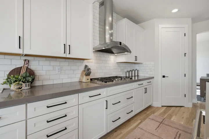 White cabinetry with matte black hardware, subway tile backsplash, dark-toned countertops, stainless steel range hood, and LVP wood-finish flooring.