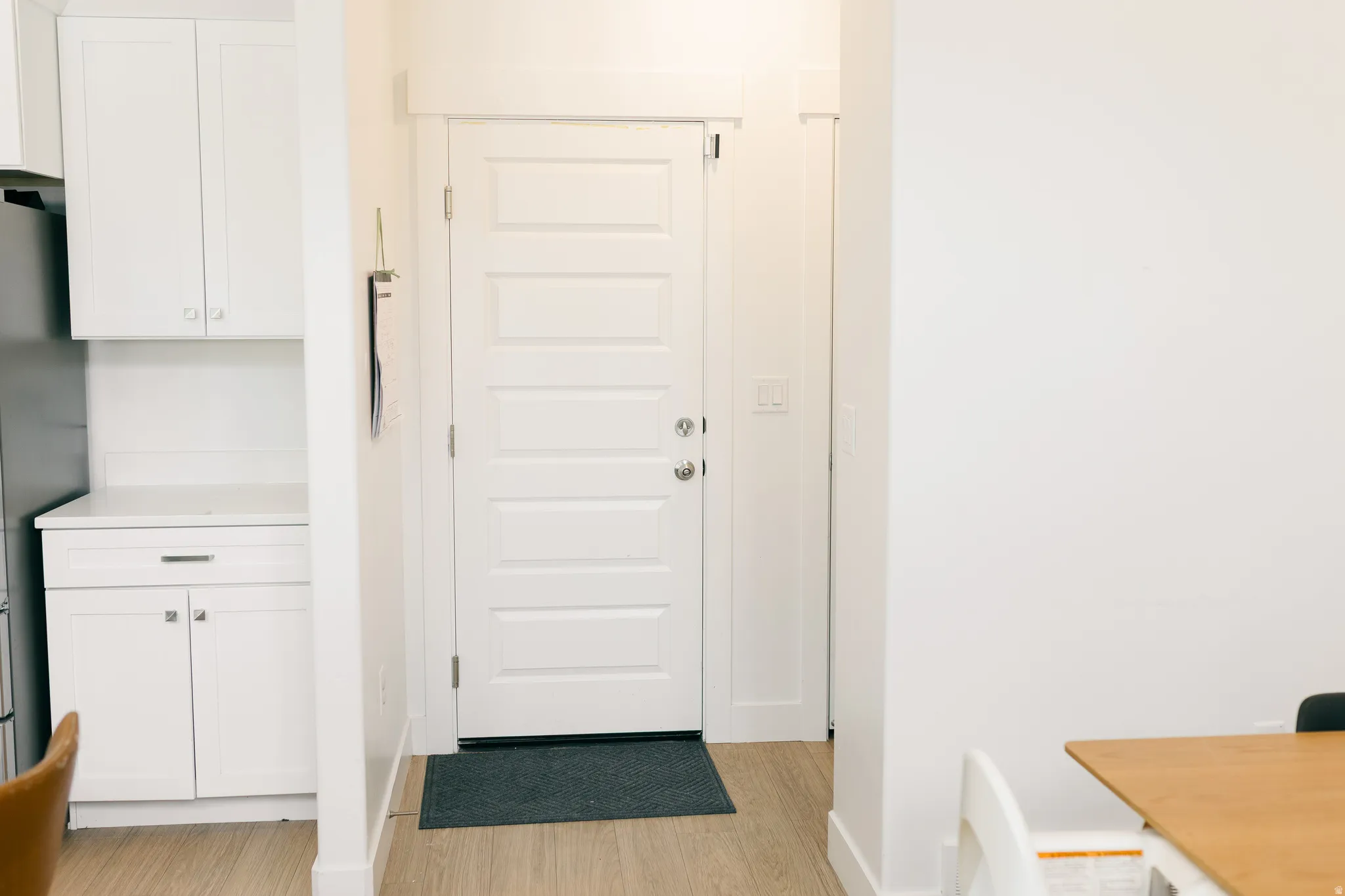 White paneled entry door with brushed nickel hardware, white shaker-style cabinetry, light wood-finish flooring, and white walls