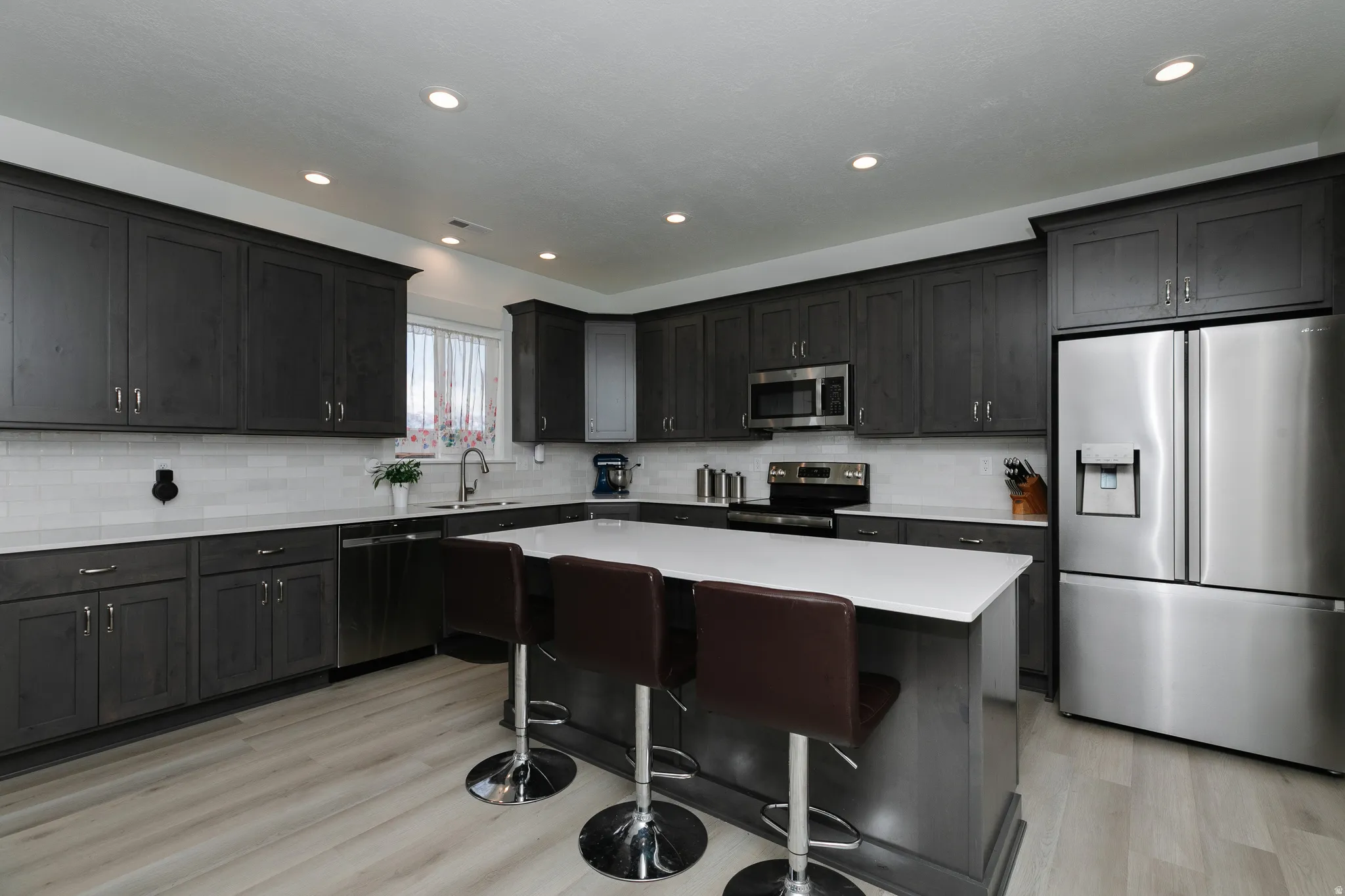 Modern kitchen featuring dark gray cabinetry, white countertops, stainless steel appliances, a white subway tile backsplash, and light wood-finish flooring