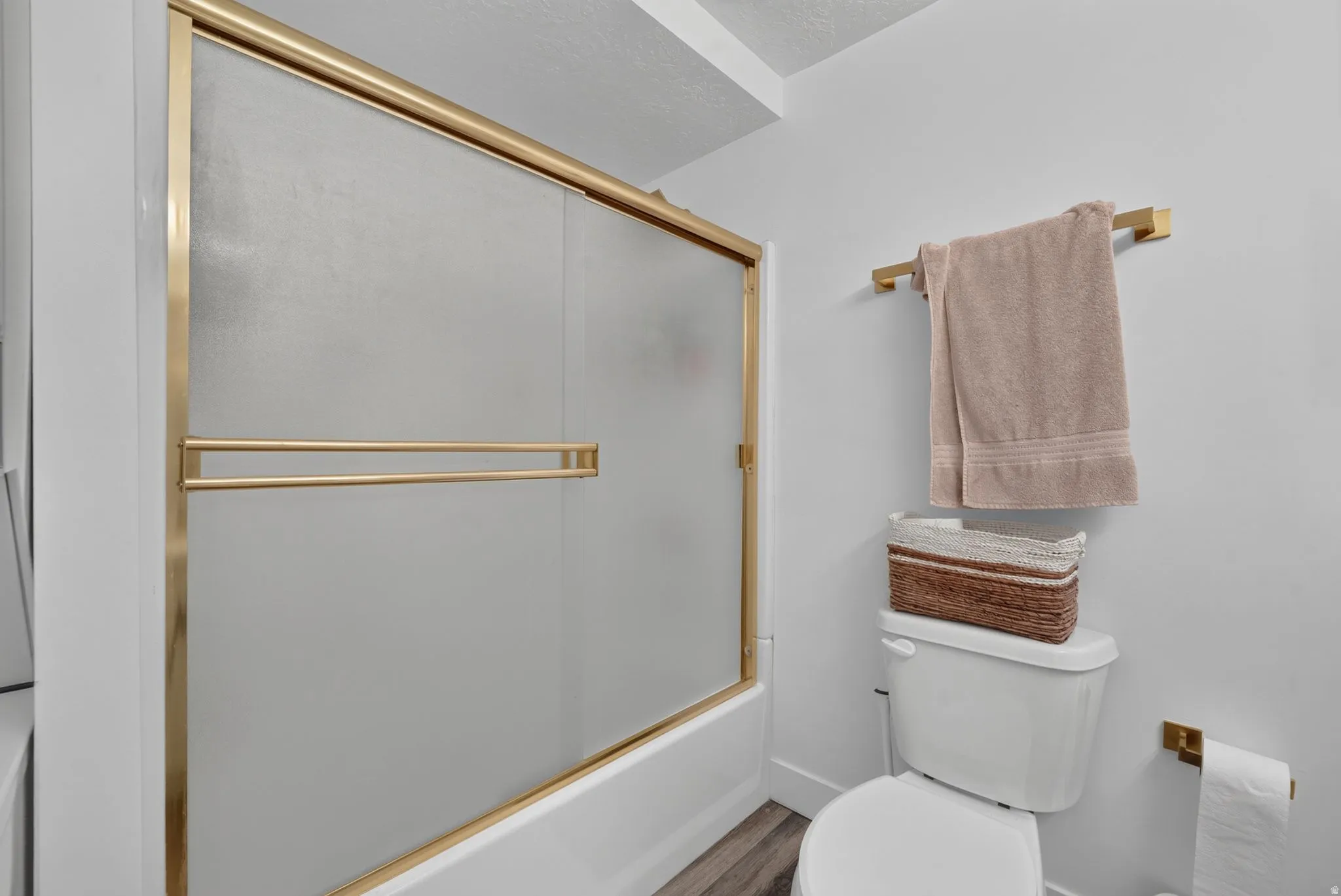 White bathtub with brass-finish sliding glass enclosure, white toilet, brass-finish towel bar, and wood-finish flooring